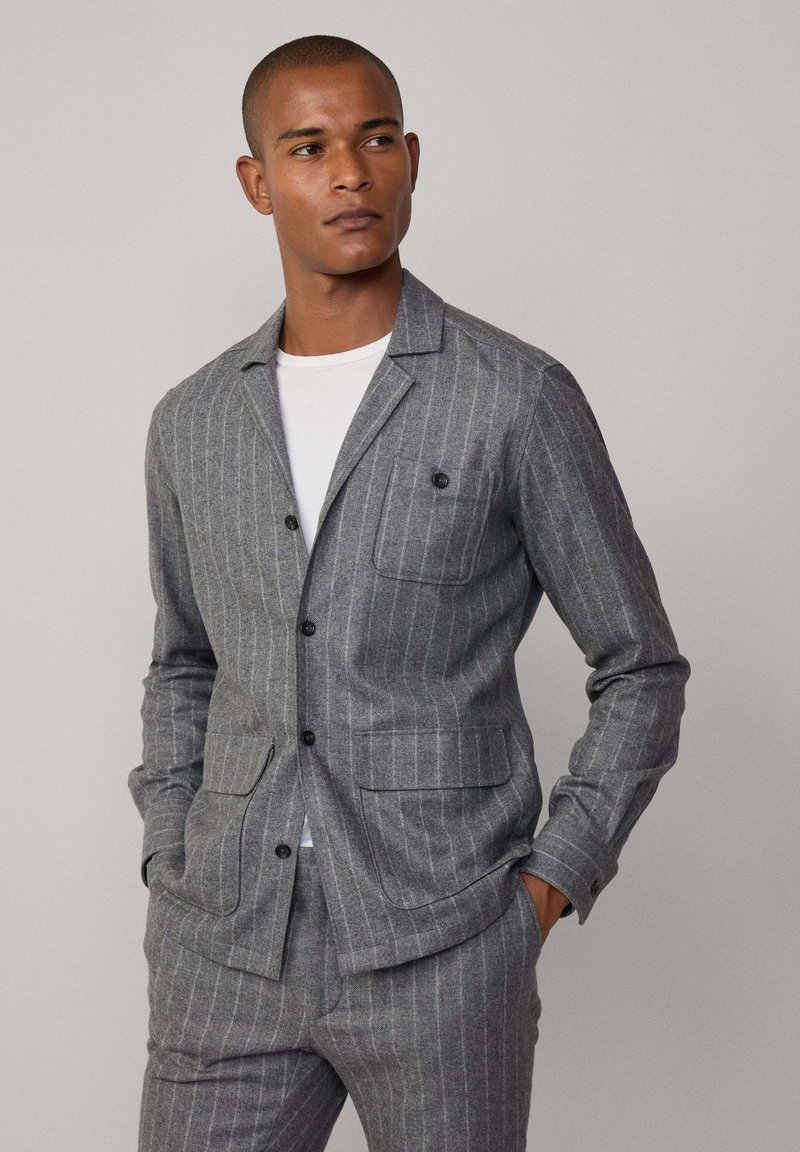 Giacca slim-fit grigia a righe sottili realizzata in tessuto texture, con colletto a spacco, due tasche frontali e chiusura a bottoni. Indossata sopra una camicia bianca.