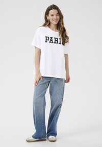 T-shirt en coton blanc avec un texte "PARIS" noir en gras, associé à un jean en denim bleu clair à jambes larges et des baskets beiges avec des accents blancs.
