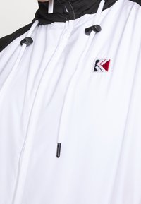 Giacca a vento bianca con accenti neri, dotata di lacci regolabili, colletto alto e un piccolo logo multicolore. Materiale morbido e leggero.