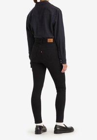 Jeans skinny negros con cintura alta, bolsillos traseros en forma de corazón y un parche de cuero marrón claro en la cintura. Llevados con una camisa oscura y zapatos negros.