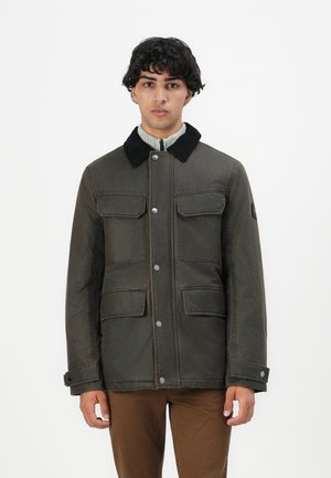 Junger Mann in einer dunkelgrünen Jacke mit schwarzem Kragen und vier Klapptaschen, kombiniert mit braunen Hosen, steht vor einem weißen Hintergrund.