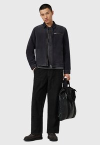 Giubbotto di pelle nera, t-shirt grigia, pantaloni di velluto a coste neri e scarpe nere. Tiene una borsa nera testurizzata, in piedi contro uno sfondo neutro.