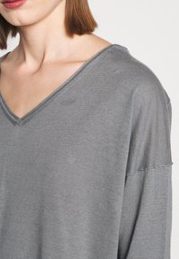 Femme portant un pull gris ample à col en V avec des épaules tombantes, dévoilant une partie du cou et de la mâchoire sur un fond blanc.