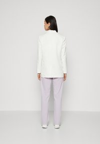 Blazer bianco a righe sottili con maniche lunghe, abbinato a pantaloni lilla chiaro. L'outfit presenta una vestibilità su misura e accenti semplici.