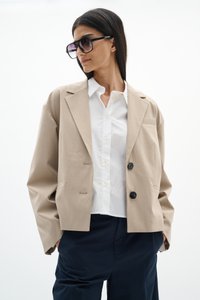 Frau trägt einen beige übergroßen Blazer, ein weißes Hemd, marineblaue Hose und große schwarze Sonnenbrille, steht mit Händen in den Taschen.