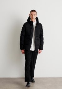 Geacă puffer neagră, cu închidere cu fermoar, guler înalt și două buzunare frontale, purtată cu o cămașă albă și pantaloni negri. Textură netedă.