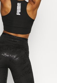 Svart sport-bh med vitt "PUMA"-logotyp på baksidan, kombinerad med svarta leggings med ett glansigt, texturerat mönster.