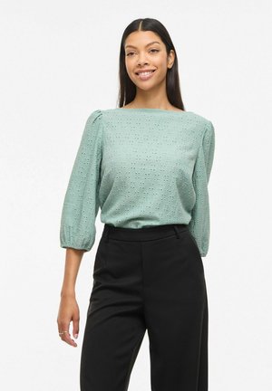 Kvinde smilende iført en lysegrøn bluse med hulmønster og trekvartlange puffærmer samt sorte bukser med høj talje mod en hvid baggrund.