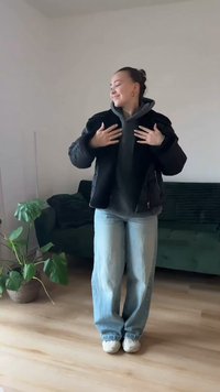 Čierna nadýchaná bunda cez sivú mikinu s kapucňou, doplnená svetlomodrými širokými džínsami. Prostredie zahŕňa zelenú pohovku a kvetináč s rastlinou.