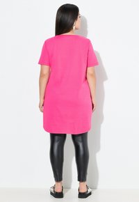 Roze katoenen T-shirt jurk met korte mouwen, vallend onder de knieën, gecombineerd met zwarte glanzende leggings en zwarte instap schoenen.