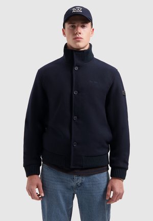 Marineblauwe collared jacket met knoopsluiting, ribgebreide mouwen en zoom, textuurstof en logo-detail op de borst. Gekleed met lichte jeans.