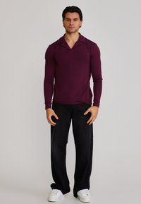 Bărbat îmbrăcat cu un tricou polo bordo cu mânecă lungă, pantaloni negri evazați și adidași albi, stând în picioare pe un fundal simplu.