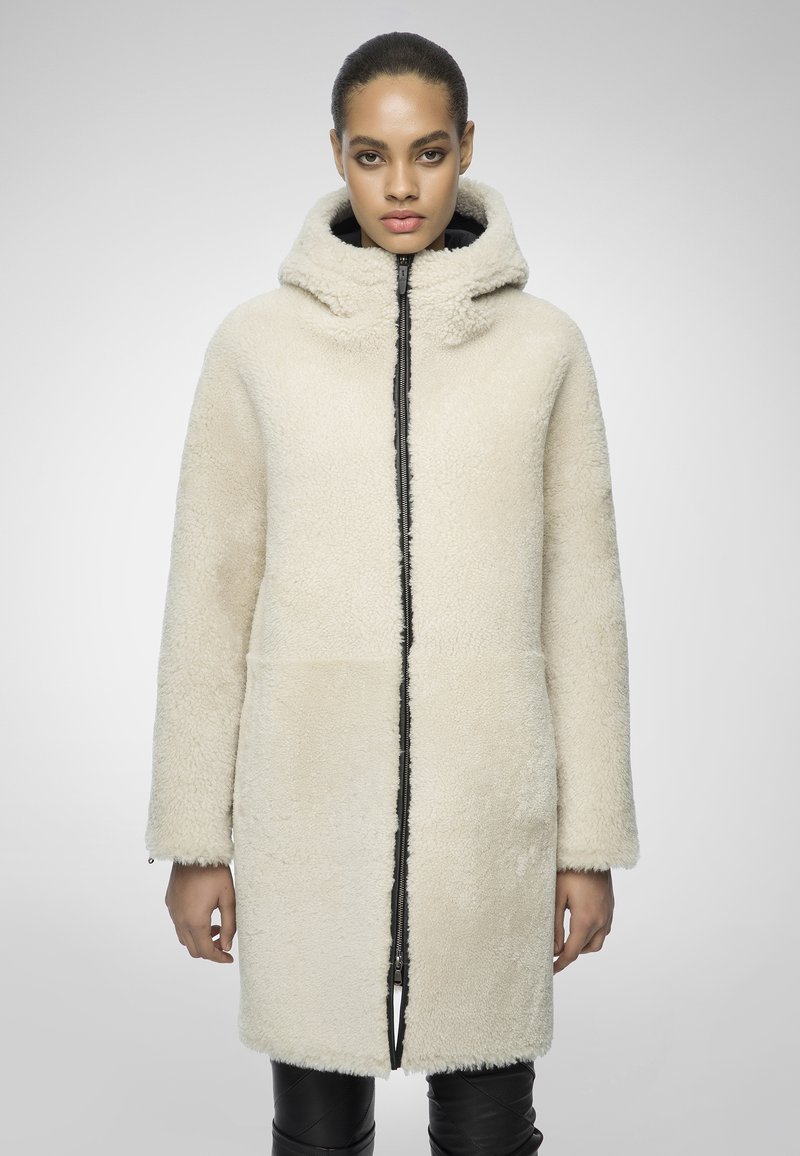 Donna in piedi che indossa un lungo cappotto fleece color crema con cappuccio e zip frontale, abbinato a pantaloni neri, su uno sfondo chiaro e semplice.