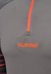 Grijs sportshirt met oranje rits en de merknaam "hummel" in het oranje gedrukt op de borst.