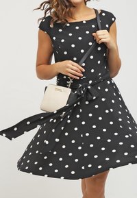 Mustapuku, jossa on valkoisia pilkkuja, hihat, vyötärö sidottuna rusetiksi, yhdistettynä pieneen beigekuosiseen käsilaukkuun, jossa on ketjusolka.