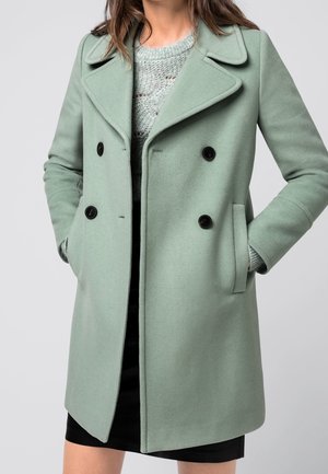 Femme portant un manteau en laine vert clair à double boutonnage par-dessus un pull en tricot gris et une jupe noire, les mains dans les poches du manteau, sur un fond uni.