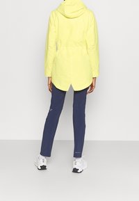 Ljusgul jacka med huva och elastisk midja, i kombination med marinblå byxor. Vita träningsskor fulländar outfiten.