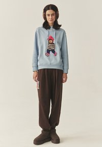 Világoskék kapucnis pulcsi színes medve hímzéssel, barna bőszárú nadrággal és barna szőrös csizmával párosítva. A nadrágon színes folt található.