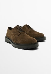 Chaussures en suède marron avec un bout arrondi, un profil bas et une surface texturée. Elles sont dotées de lacets noirs et d'une semelle en caoutchouc épaisse et robuste.