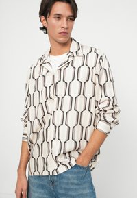 Camicia a maniche lunghe in un tessuto morbido e leggero con un motivo geometrico nero e beige. Presenta una vestibilità ampia e un colletto con bottoni.