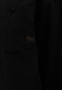 Camisa de ganga preta com frente de botões e bolso no peito, com um pequeno rótulo de tecido que diz "G-STAR RAW". Superfície texturizada.