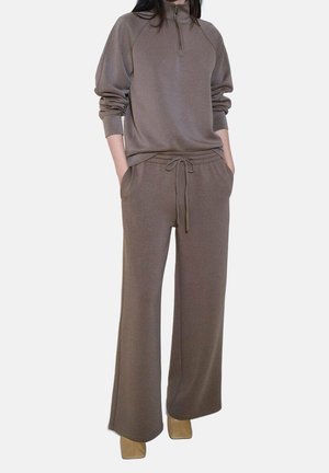 Femme portant un pantalon en maille marron ample avec taille à cordon et un pull à manches longues assorti avec demi-fermeture éclair, mains dans les poches, chaussures beige visibles.