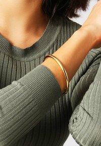 Bracciale rigido d'oro indossato su un polso, con una finitura metallica liscia, posato su un maglione verde oliva testurizzato con dettagli a coste.
