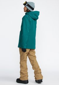 Veste imperméable turquois avec capuche, gants noirs, pantalon de neige beige et bottes noires. Modèle de profil droit sur fond blanc uni.