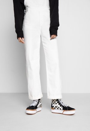 Person iført hvide overaller og sort-hvide ternede høje platform sneakers, stående mod en ensfarvet lys baggrund.