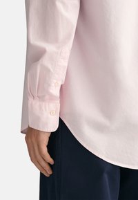 Helles rosa Langarmshirt mit geknöpften Manschetten, das eine glatte Textur und einen abgerundeten Saum aufweist, kombiniert mit dunkelblauen Hosen.