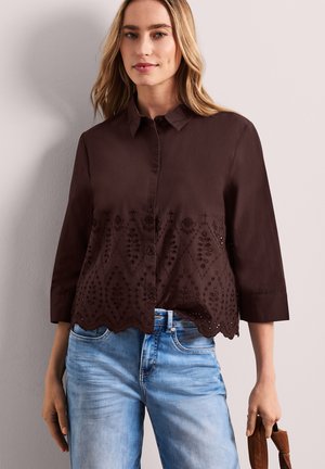 Mujer que lleva una blusa marrón oscuro con botones, con bordados florales y dobladillo con ondas, combinada con vaqueros azul claro, sosteniendo un bolso marrón.