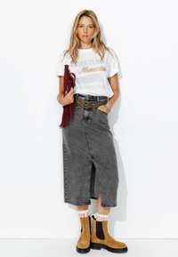 Jupe midi en denim gris avec fente avant, associée à un t-shirt graphique blanc et des bottines en suède marron. Sac à franges rouge et chaussettes à motifs inclus.