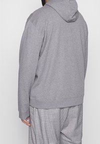 Personne portant un sweat à capuche gris uni et un pantalon ample gris à carreaux, vue de dos sur un fond neutre.