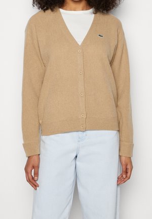 Kvinde iført en beige knappet cardigan over en hvid skjorte og lyseblå jeans, stående foran en ensfarvet baggrund.