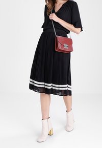Gonna nera plissettata con strisce bianche all'orlo, abbinata a una borsa a tracolla in pelle rossa e stivaletti beige con tacco dorato.