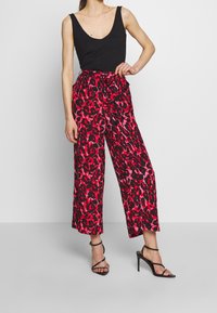 Pantalons large à imprimé léopard rouge et noir avec taille haute et ceinture en tissu, accompagnés d'un top sans manches noir et de sandales à talons noires.