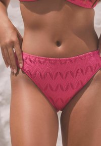 Rosa Bikini-Höschen mit strukturiertem Stoff und wellenförmigen Mustern, mittelhohem Schnitt und glatten Kanten, hervorgehoben auf gebräunter Haut.