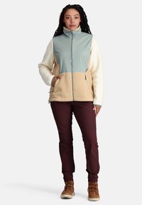 Fleecepullover und Softshelljacke in Hellgrün, Creme und Beige mit Reißverschlusstaschen; kombiniert mit burgunderfarbenen Hosen und braunen Wanderschuhen.