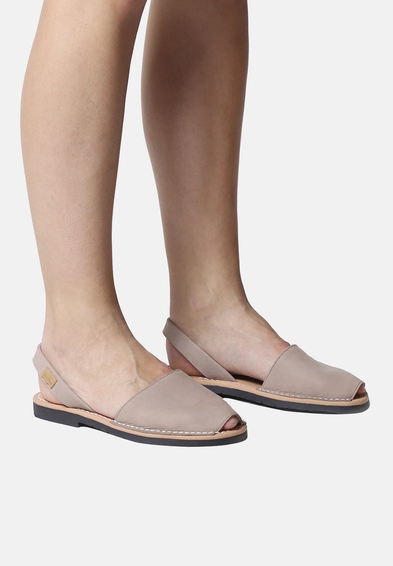 Paire de sandales plates beiges à bout ouvert avec bride arrière, portées sur des pieds avec des tons de peau clairs et moyens, sur un fond uni.