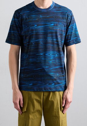 Mężczyzna w granatowo-czarnej, wzorzystej koszulce z krótkim rękawem z motywem słojów drewna oraz oliwkowych szortach typu cargo.