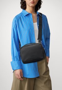 Svart crossbody-väska med slät textur, rektangulär form, justerbar axelrem och diskret logotyp, bärs över en blå randig skjorta.