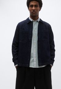 Σκούρο τζιν μπουφάν με δύο μπροστινές τσέπες, φορεμένο πάνω από ένα ανοιχτόχρωμο ριγέ πουκάμισο με κουμπιά. Το μοντέλο στέκεται με τα χέρια στις τσέπες, φοράει μαύρο παντελόνι.