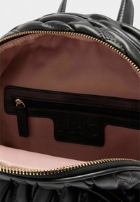 Bolso de cuero acolchado negro abierto para mostrar un interior de tela suave color rosa y un bolsillo negro con cremallera y tirador dorado.
