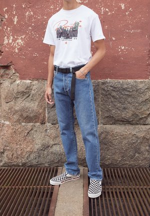 T-shirt graphique blanc avec un design en vert et rouge, assorti à un jean bleu clair et une ceinture noire. Les chaussures présentent un motif à carreaux.