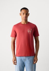 Moški nosi rdečo majico s kratkimi rokavi z majhnim naravnim motivom na levi prsni strani in svetlo modre traper hlače, stoji ob belem ozadju.