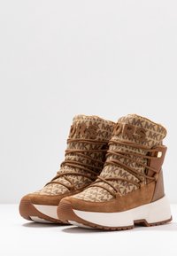 Bottines en daim marron et tissu à motifs avec de épaisses semelles blanches, présentant des détails de logo sur la tige et un design à lacets.