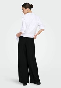 Bílá košile s límečkem a zahnutými rukávy, v kombinaci s černými, širokými kalhotami s proužky. Má jednoduchý, čistý design a hladkou texturu.