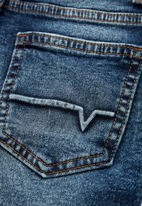 Bolsillo trasero de unos jeans de mezclilla con tela azul, costuras naranjas y un distintivo patrón de zigzag blanco bordado en el bolsillo.