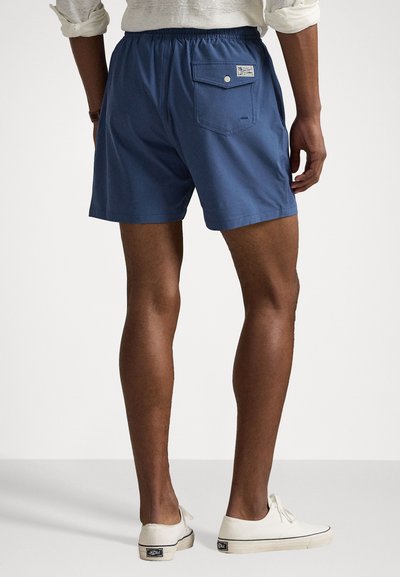 Blue swim shorts with an elastic waistband, featuring a back pocket with a button closure. Worn with white canvas shoes.
