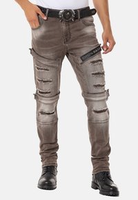 Abgedrehte graue Denim-Jeans mit mehreren ausgefransten Details, tailliertem Design und einem schwarzen Gürtel mit Totenkopf-Motiv und silberner Hardware.